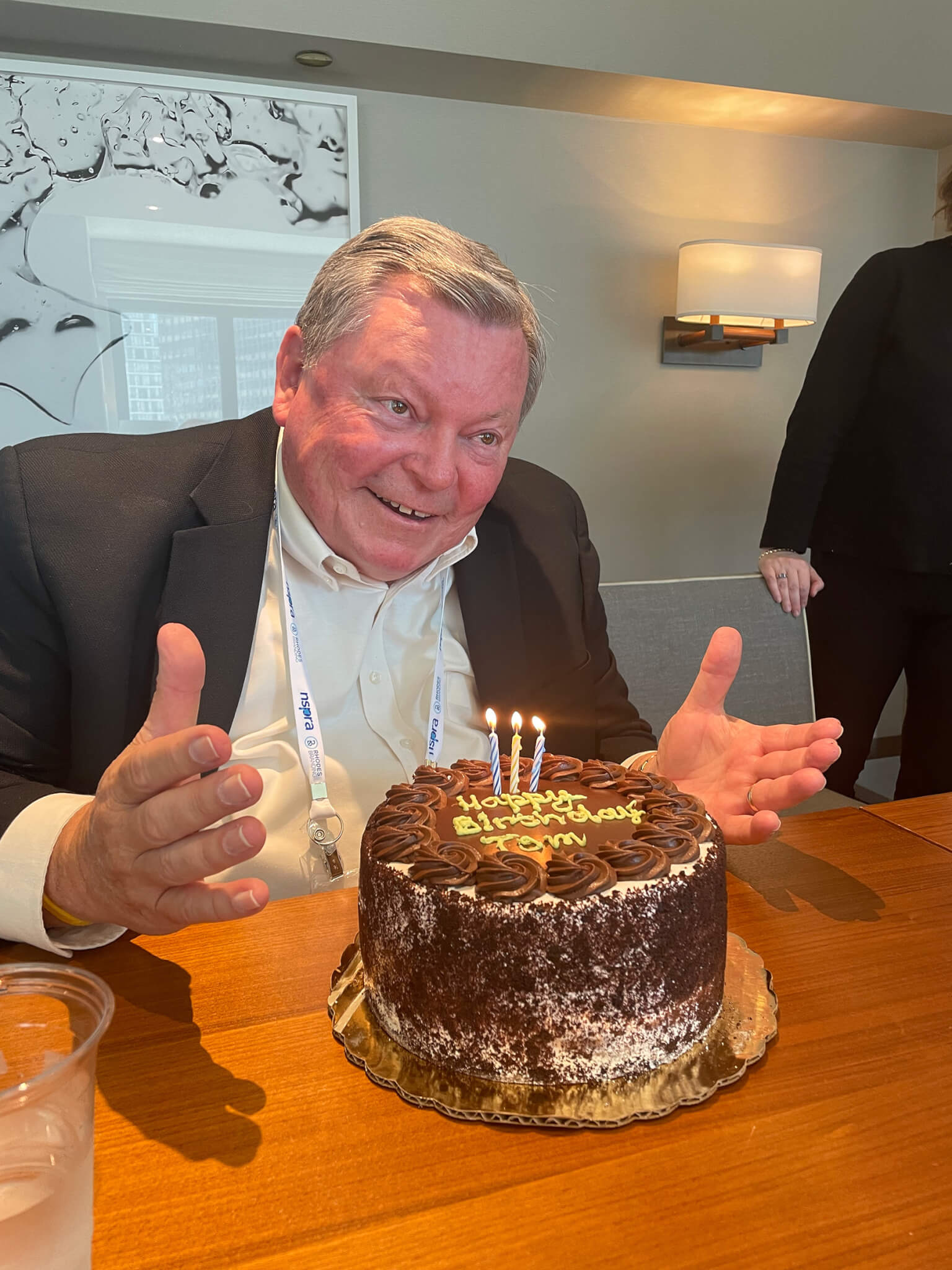 A man in business dress smiles and gestures while seated in front of a birthday cake with three lit candles, covered in chocolate frosting. The cake reads Happy Birthday Tom