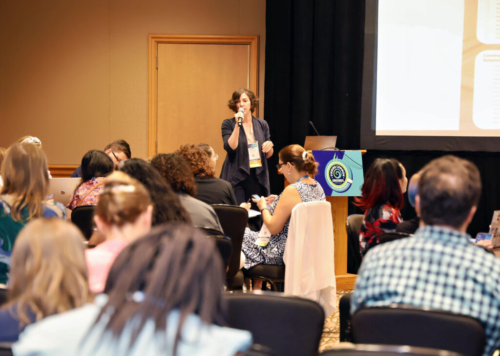 A white woman spekas into a microphone in a room crowded full of seated people